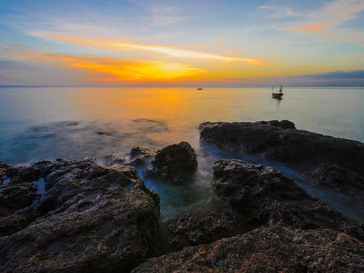 Sonnenuntergang über ruhigem Wasser, Felsen im Vordergrund und ein Boot entfernt auf dem Meer.